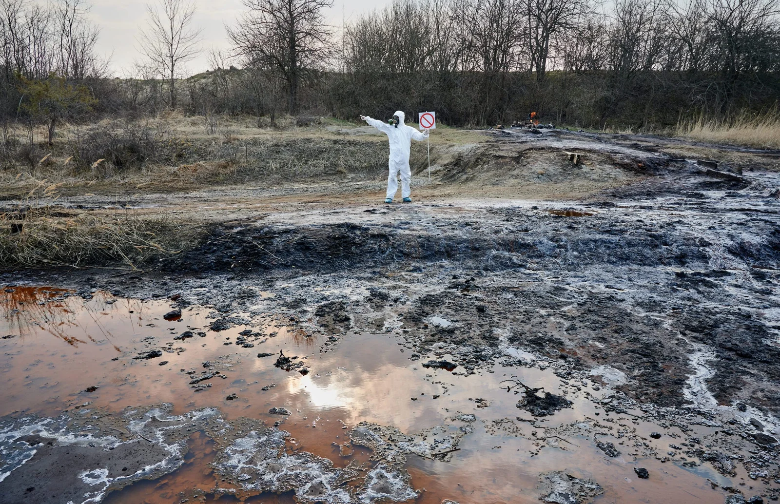 Man in white PPE holding a "no" icon sign standing near what looks like contaminated ground